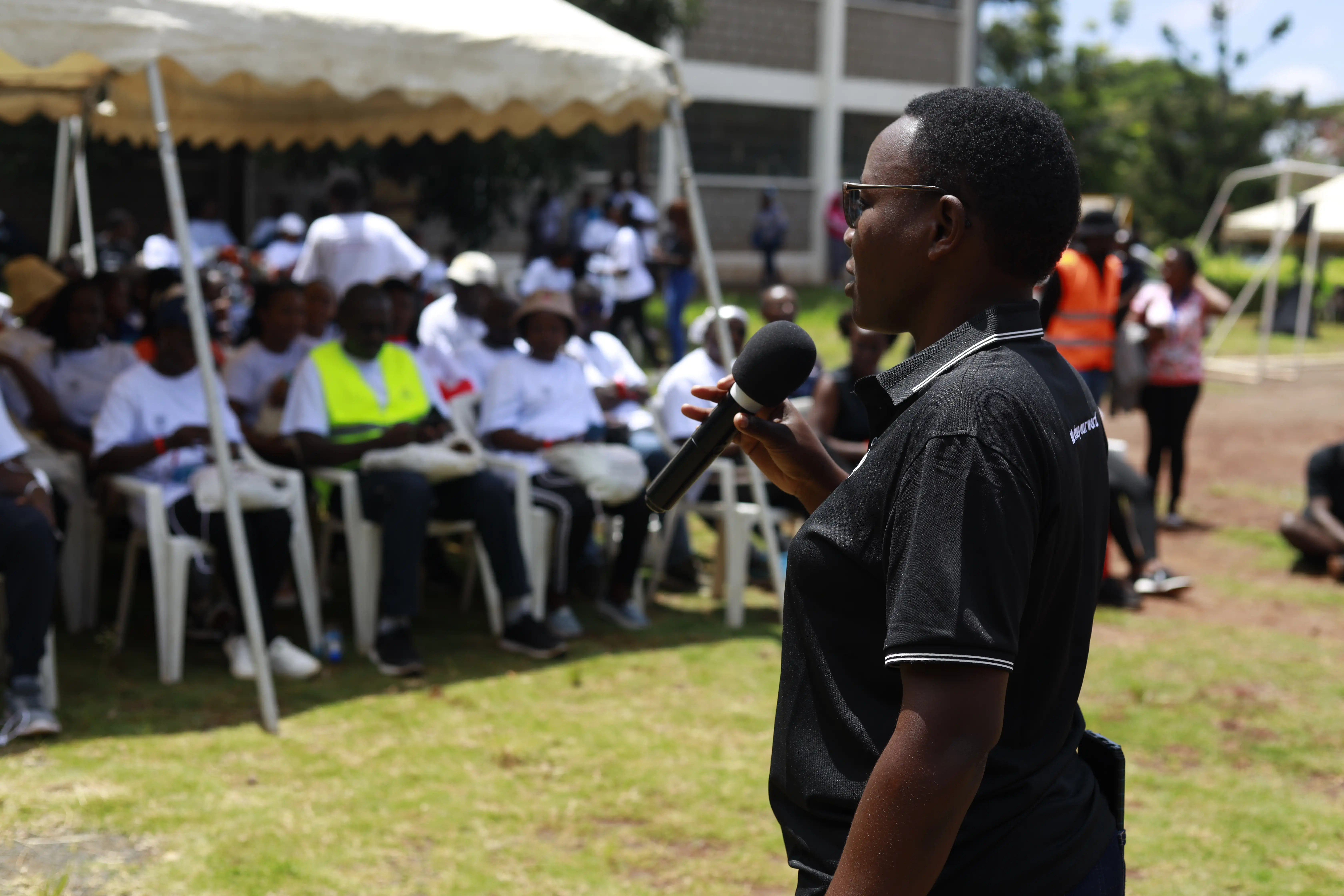 Man in KCB Club t-shirt speaking into a microphone outdoors